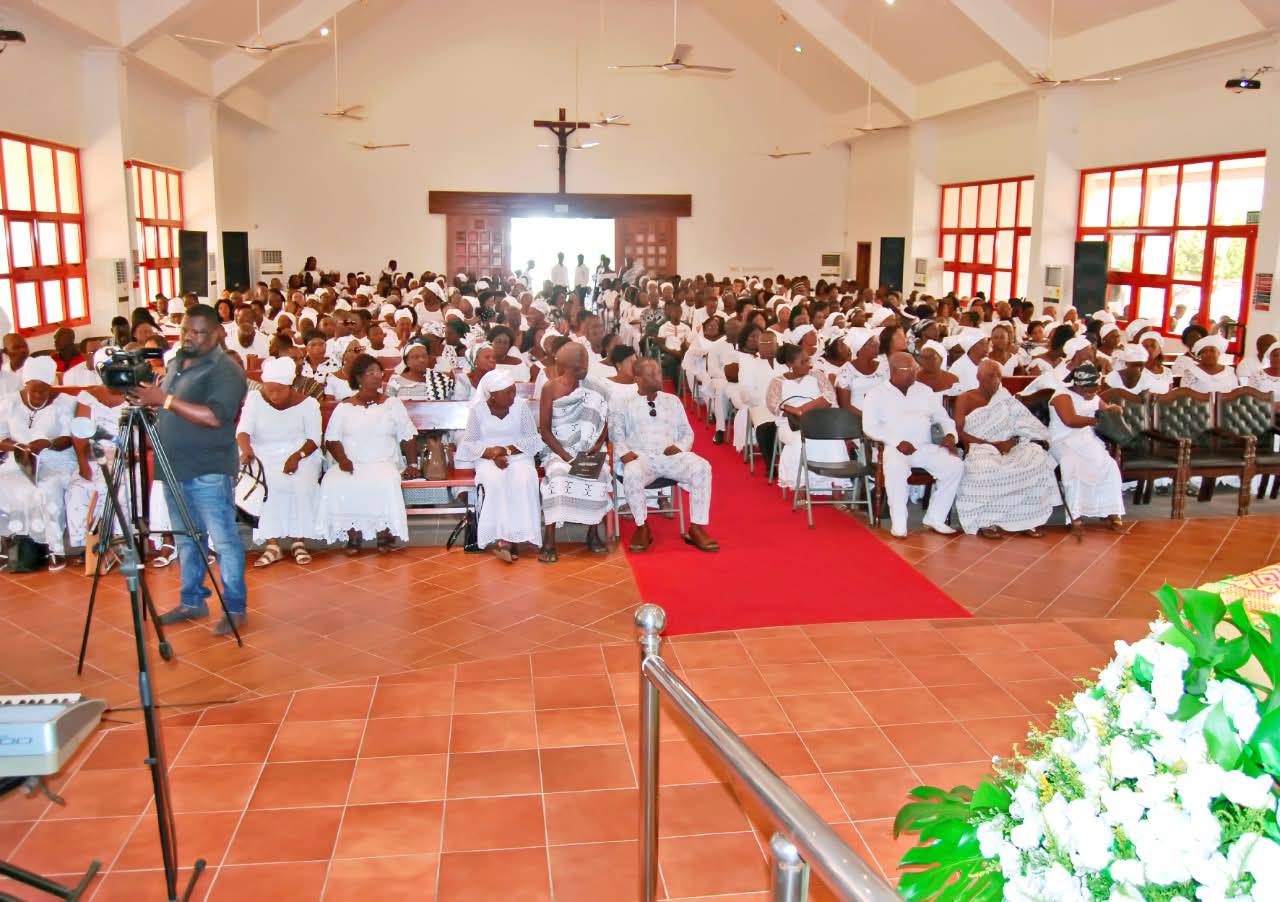 An expansive view from the sanctuary platform capturing the congregation seated in traditional white funeral attire along the central red aisle runner.